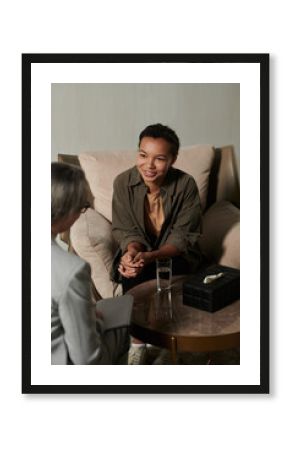 Happy young African American female patient sitting in comfortable armchair in front of psychologist and looking at her during session