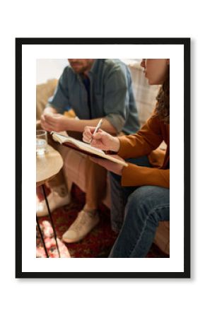 Vertical image of therapist talking to patient and making notes in notepad while they sitting on sofa in office