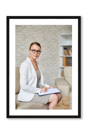 Portrait of beautiful female psychologist wearing glasses posing with clipboard in therapy office looking to camera