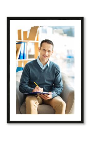 Professional psychologist. Cheerful happy nice man sitting in the armchair and taking notes while working as a psychologist