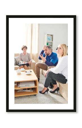 Angry emotional bearded husband sitting on sofa and pulling hair on head while screaming at wife and gesturing hand during therapy session with psychologist
