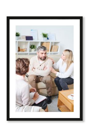 Serious pensive middle-aged husband with gray hair sitting on sofa and being distant while listening to pretty wife at therapy session, young woman touching his shoulder and supporting him