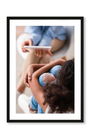 Overhead view of african american girl hugging legs during consultation with blurred psychologist on background