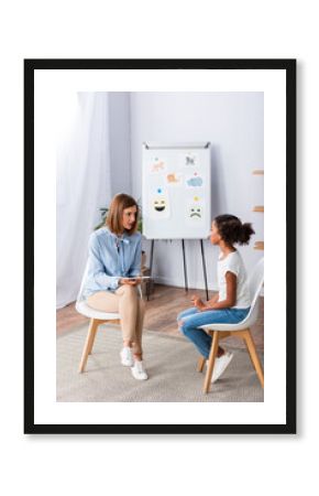 Full length of female psychologist with digital tablet talking to african american girl near flipchart in office