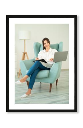 Young woman sits in chair with laptop, looks camera and holds pen in her hands.