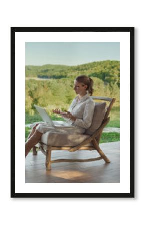 Woman sitting outdoors on terrace with a laptop, engaged in an online video meeting or counseling, discussing remotely in a calm, natural environment surrounded by greenery. 