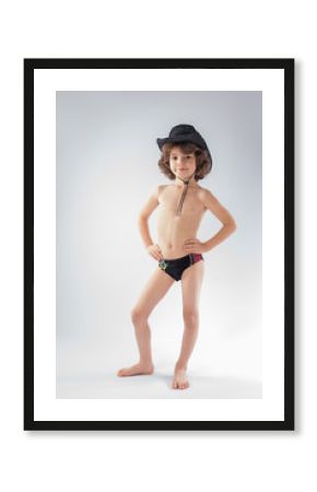 The curly-haired boy in a bathing suit and cowboy hat is looking into the camera and smiling. Gray background.