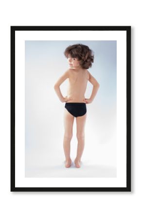 Curly boy in swimming trunks standing in full length with his back to the camera and looking into the camera. Gray background.