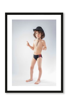 Little curly-haired cowboy hat and bathing suit standing and looking at the camera. Gray background.