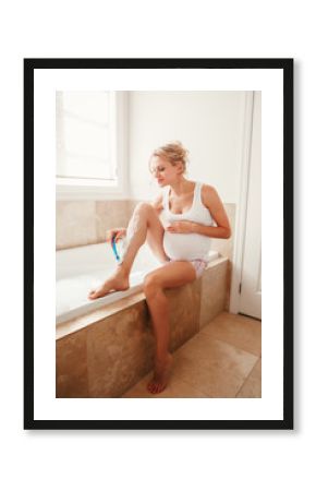 Problems and difficulties during pregnancy. Portrait of pregnant blonde Caucasian woman shaving legs in bathroom. Expecting mom doing cosmetological procedures in the morning.