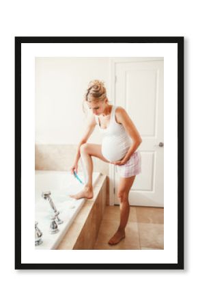 Problems and difficulties during pregnancy. Portrait of pregnant blonde Caucasian woman shaving legs in bathroom. Expecting mom doing cosmetological procedures in the morning.