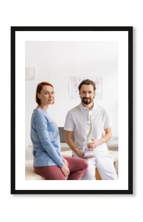 happy redhead woman and smiling physiotherapist with spine model looking at camera in consulting room.