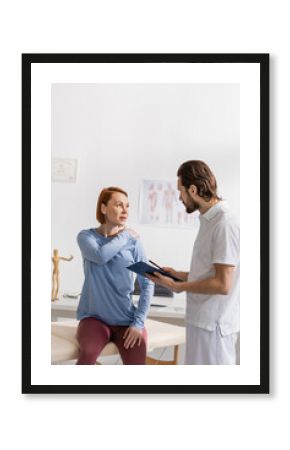 woman touching painful shoulder during consultation with physiotherapist holding clipboard in consulting room.