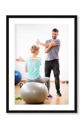 Middle age blond woman exercising on gymnastic ball during session with physiotherapist