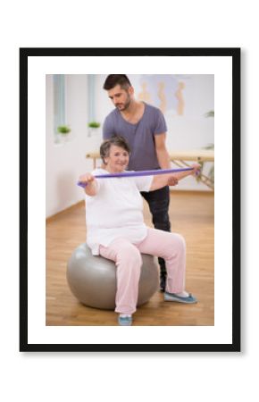 Professional physiotherapist stabilizing senior woman sitting on exercising ball