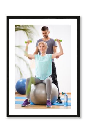 Middle age blond woman exercising on gymnastic ball during session with physiotherapist