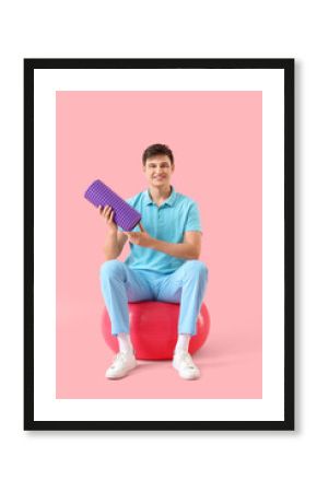 Male physiotherapist with foam roller sitting on fitball against pink background