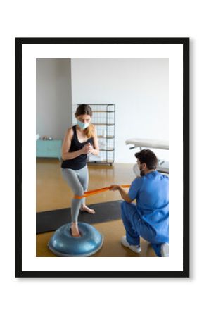 Woman in face mask doing squats using resistance elastic rubber band for rehabilitation exercise for legs, with physiotherapist or doctor assisting her in a health clinic during Coronavirus outbreak.