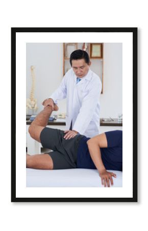 Vertical medium long shot of mature Asian physical therapist wearing white coat working with unrecognizable male patient recovering his hip joints range of motion