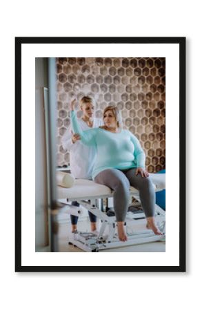 Mid adult physiotherapist woman exercising with overweight woman indoors in rehabilitation center