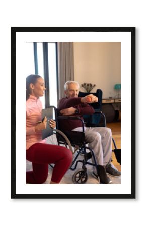 Smiling biracial female physiotherapist looking at caucasian senior man lifting dumbbell at home