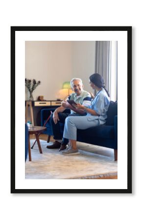 Biracial female physiotherapist using digital tablet while talking with caucasian senior man on sofa