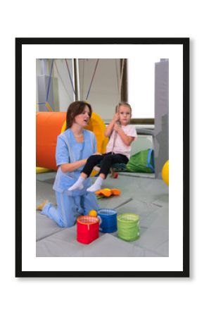 Child with physiotherapist on swing during sensory integration session. Little girl in rehabilitation centre doing exercises for development functions of the vestibular apparatus