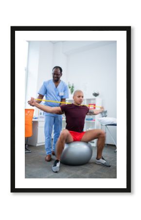 Therapist in uniform standing near man sitting on fitness ball