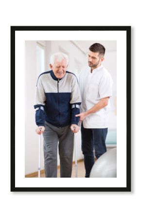 Elderly man walking on crutches and a helpful male nurse supporting him