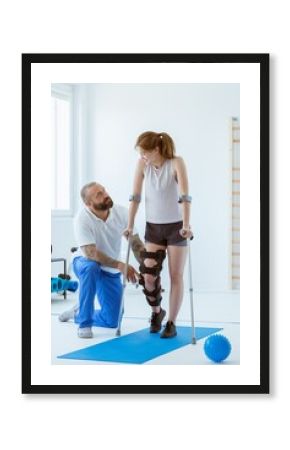 The girl after a stroke with an orthosis on a crutch leg is exercising on a blue mat in a gym in a hospital