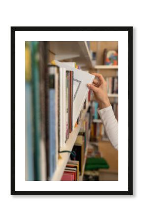 Female hand pulling book from bookshelf in public library in university, college or high school. Woman student take novel from bookcase in bookshop store, soft focus. Education and literature concept