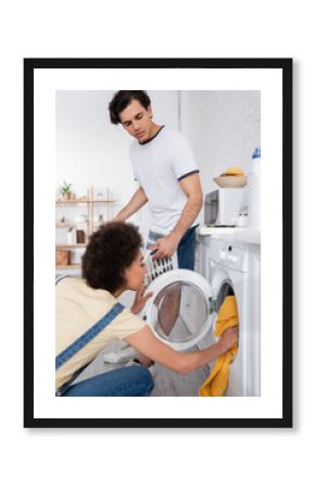 curly african american woman loading washing machine near boyfriend with basket with dirty laundry