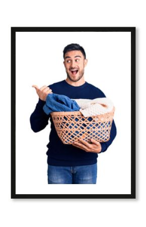 Young handsome man holding laundry basket pointing thumb up to the side smiling happy with open mouth