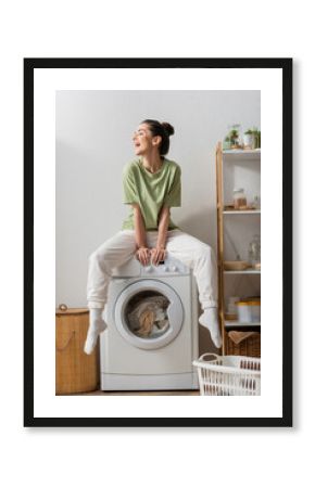 Excited young woman sitting on washing machine in laundry room.