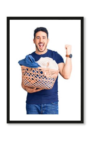 Young hispanic man holding laundry basket screaming proud, celebrating victory and success very excited with raised arms