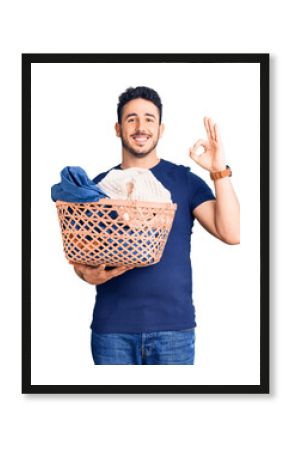 Young hispanic man holding laundry basket doing ok sign with fingers, smiling friendly gesturing excellent symbol