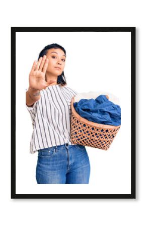 Young woman holding laundry basket with open hand doing stop sign with serious and confident expression, defense gesture