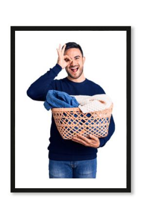 Young handsome man holding laundry basket smiling happy doing ok sign with hand on eye looking through fingers