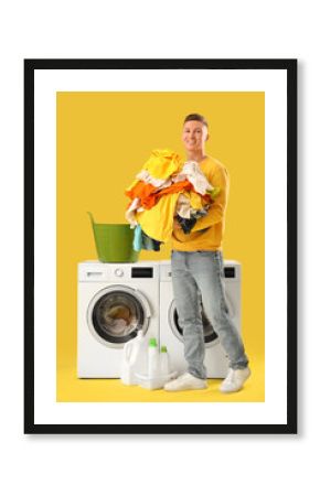 Handsome young man holding pile of laundry near washing machine on yellow background