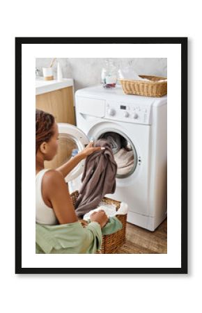 An African American woman with afro braids carefully loads clothes into a washing machine in a bathroom.