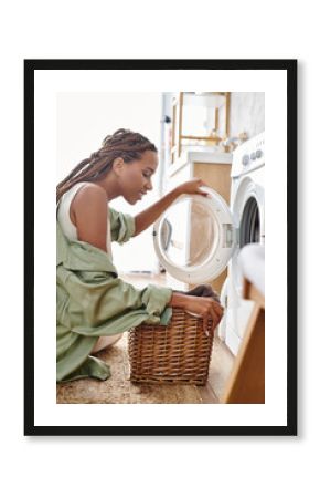 An African American woman with afro braids sits next to a washing machine in a bathroom, focused on doing the laundry.
