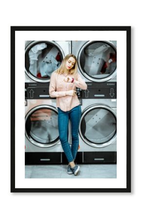 Young woman standing near the dryer machines waiting for clothes to be dried in the self serviced laundry