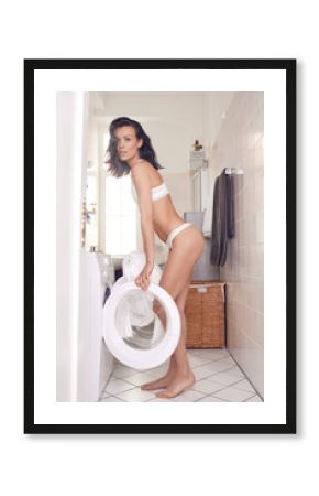 Sexy attractive middle-aged woman in white underwear filling the washing machine standing in front of the open door in her bare feet in a tiled laundry in a side view