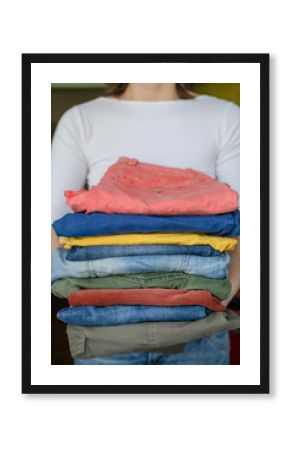 Woman holds neatly folded trousers and jeans in bright colors, after washing, in the laundry room.