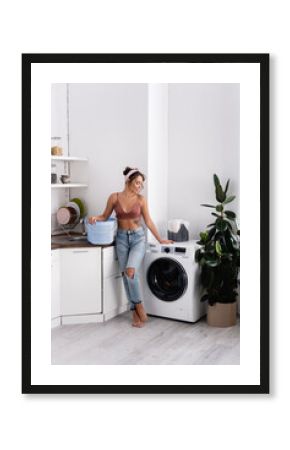 Barefoot housewife holding laundry basket near washing machine in kitchen