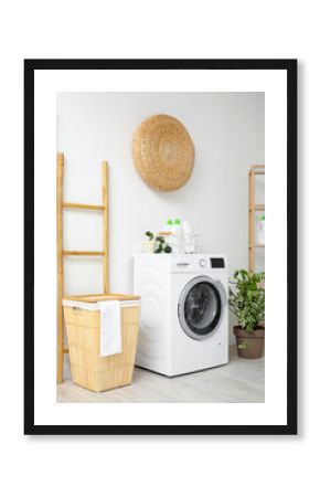 Modern washing machine with basket, houseplant and ladder near white wall. Interior of home laundry room