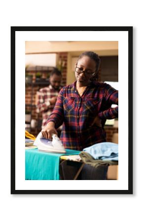 Black woman doing household chores looking tired while ironing clothes in living room. Young female adult holding waist, showing signs of fatigue and body tension at home during weekend chores.