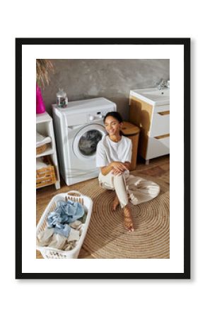 In a modern apartment, a young african american woman relaxes while doing laundry, smiling.