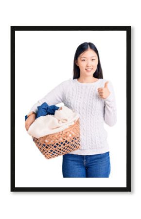 Young beautiful chinese woman holding laundry basket with clothes smiling happy and positive, thumb up doing excellent and approval sign