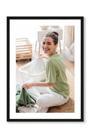 Cheerful young woman looking at camera near clothes and basket on floor in laundry room.